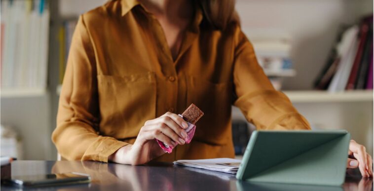 An anonymous businesswoman sitting at the office, eating a protein bar while video calling with her colleagues.