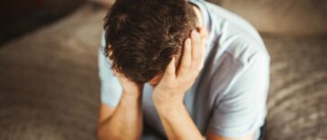 Sad man sitting on a side of the bed, holding his head in both hands.