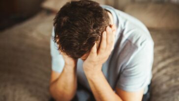 Sad man sitting on a side of the bed, holding his head in both hands.