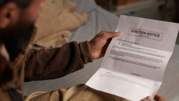 Person holding eviction notice while sitting on couch and reading in a dimly lit room without natural light coming in through the window behind him.