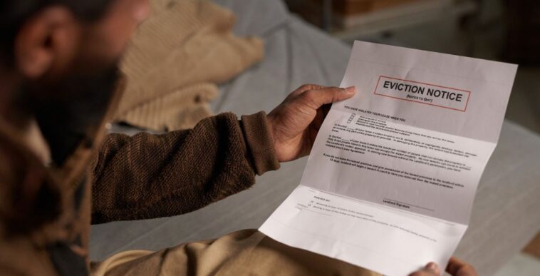 Person holding eviction notice while sitting on couch and reading in a dimly lit room without natural light coming in through the window behind him.