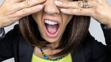 A funny fisheye image of a female with brunette hair trying to cover her eyes as she peeks though her fingers to look at the proverbial trainwreck in front of her.
