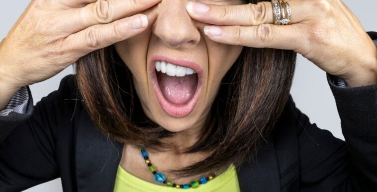 A funny fisheye image of a female with brunette hair trying to cover her eyes as she peeks though her fingers to look at the proverbial trainwreck in front of her.