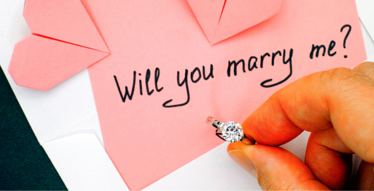 A man holding a ring in front of a pink piece of paper with "will you marry me" written on it.