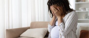 Pregnant woman sitting on a sofa at home with her face in her hands, stressed and upset, concept of anxiety, depression or emotional struggle in maternity.