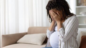 Pregnant woman sitting on a sofa at home with her face in her hands, stressed and upset, concept of anxiety, depression or emotional struggle in maternity.