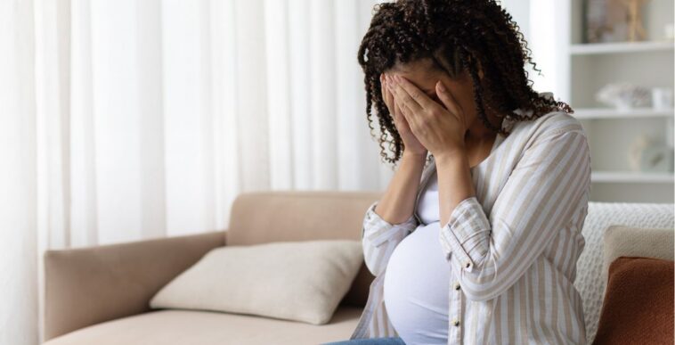 Pregnant woman sitting on a sofa at home with her face in her hands, stressed and upset, concept of anxiety, depression or emotional struggle in maternity.