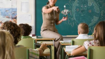 Science teacher sits on the edge of her desk, she is calling on a student and holds a light bulb.