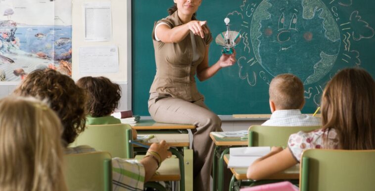 Science teacher sits on the edge of her desk, she is calling on a student and holds a light bulb.