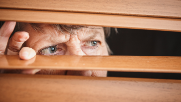 A woman peering through shades out a window.