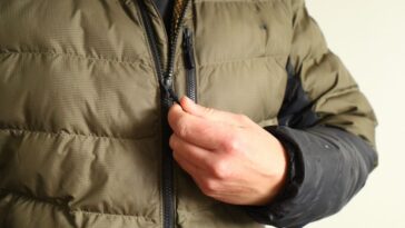 A person in a green puffer jacket zipping up, focus is on his hand and zipper, neutral background.