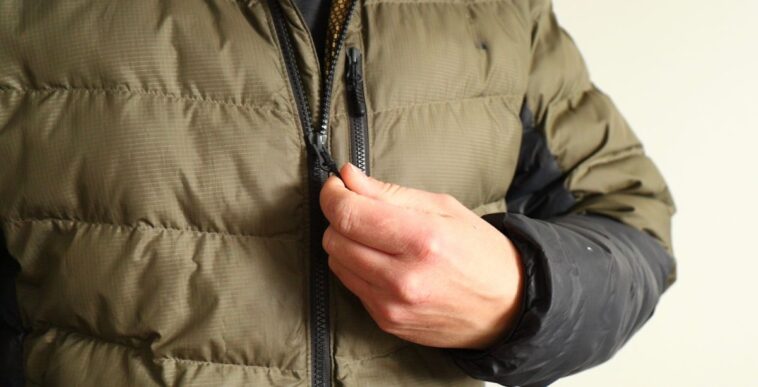 A person in a green puffer jacket zipping up, focus is on his hand and zipper, neutral background.