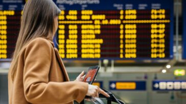 Back shot of a woman traveler checks her mobile phone in front of the airport's flight information board, preparing for her upcoming solo journey.