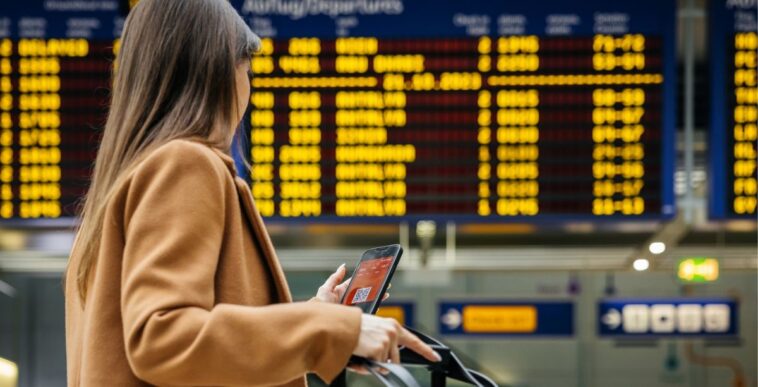 Back shot of a woman traveler checks her mobile phone in front of the airport's flight information board, preparing for her upcoming solo journey.