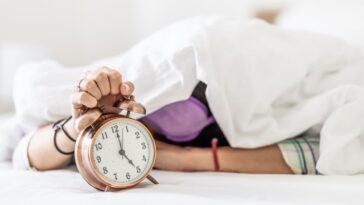 A sleepy young woman hides under the duvet as she is awakened by the loud sound of the alarm clock.