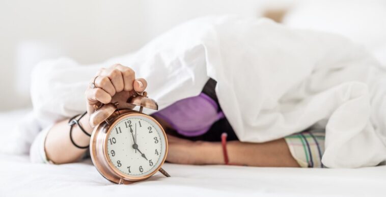 A sleepy young woman hides under the duvet as she is awakened by the loud sound of the alarm clock.