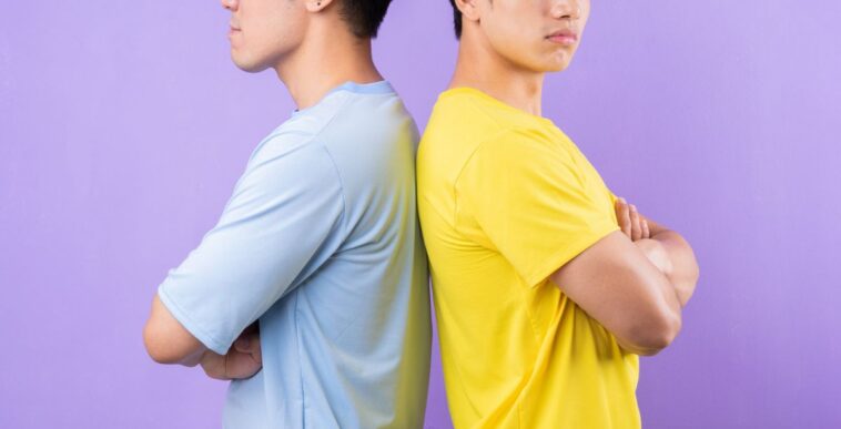 Two bothers standing back to back with their arms folded, they seem irritated. They stand against a lavender backdrop.