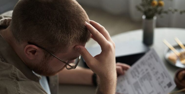 A young adult man sits at a table holding overdue bills, looking stressed and frustrated, hand on forehead, financial documents and chopsticks visible on surface.