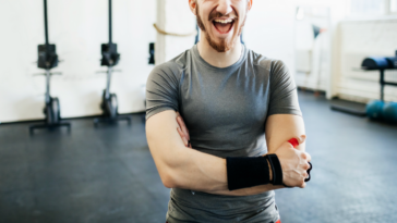 A man in a gym, folding his armns and laughing