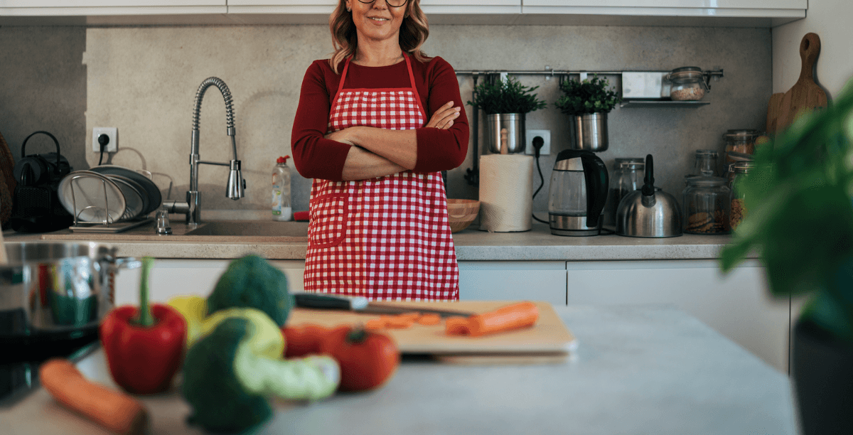 A woman wearing a red checkered apron with her arms crossed in a kitchen