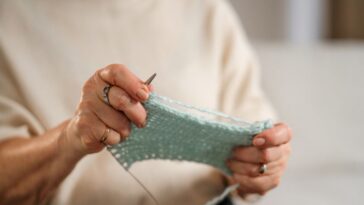 An unidentifiable senior woman sits on a comfortable sofa, knitting with focus, in a cozy, well-lit living room environment.