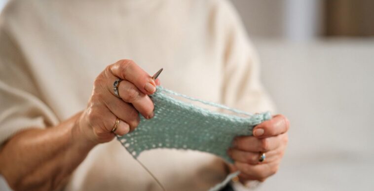 An unidentifiable senior woman sits on a comfortable sofa, knitting with focus, in a cozy, well-lit living room environment.