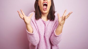 Young beautiful brunette woman wearing a pink sweater over pink isolated background crazy and mad shouting and yelling with aggressive expression and arms raised. Frustration concept.
