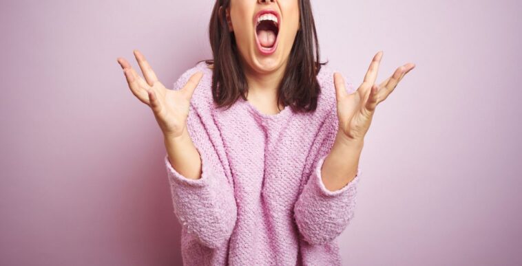 Young beautiful brunette woman wearing a pink sweater over pink isolated background crazy and mad shouting and yelling with aggressive expression and arms raised. Frustration concept.