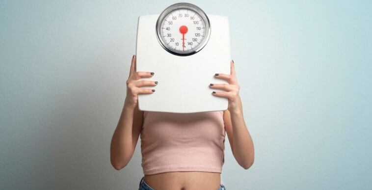 Young woman hides face behind a bathroom scale. Studio shot.