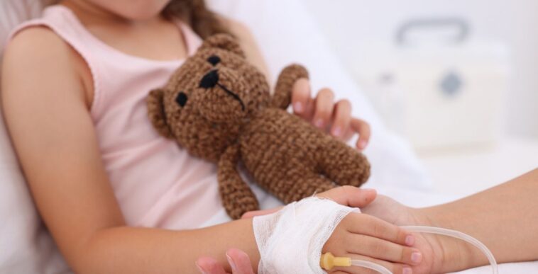Unidentifiable mother and daughter hold hands as daughter is in a hospital bed, hooked up to an IV drip. Close-up of the little girl's light brown teddy bear.
