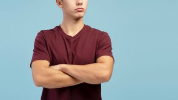 Serious, sad teen boy with crossed arms looking away on copy space, standing on blue background.