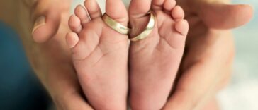 Wedding rings sit around the two big toes of a pair of baby feet while being cradled in a mother's hands.