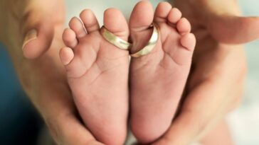 Wedding rings sit around the two big toes of a pair of baby feet while being cradled in a mother's hands.