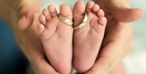 Wedding rings sit around the two big toes of a pair of baby feet while being cradled in a mother's hands.