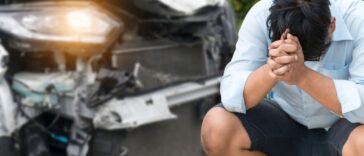 A distressed man sits on the curb in front of a car accident, he has his head in hands.