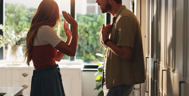 Shot of a young couple arguing in the kitchen. Woman has her head turned away and arms up defensively.