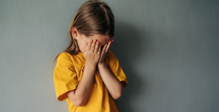 A sad, young girl cries into her hands, she stands against a gray backdrop.