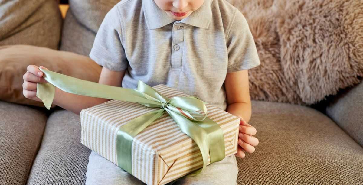 Curious young boy on a comfy sofa carefully unties a light green ribbon on a striped gift box, eagerly anticipating the surprise inside during a cozy home moment.