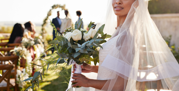 A woman wearing a bridal veil, holding a bouquet standing at the beginning of an aisle.