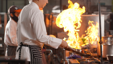 A man in a chef's coat in front of a stovetop with a big flame coming out of a frying pan.