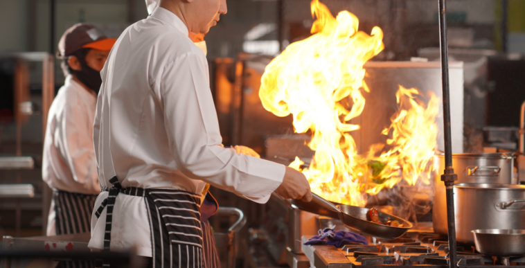 A man in a chef's coat in front of a stovetop with a big flame coming out of a frying pan.