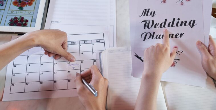 Close-up shot of unrecognizable couple wrapped up in planning wedding while sitting at table, man taking notes in calendar.