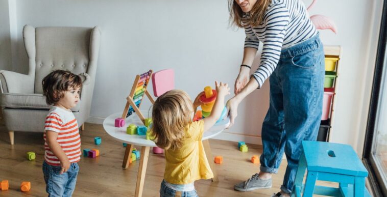 Positive young female babysitter in casual clothes playing with infant children in cozy playroom with colorful educational toys.