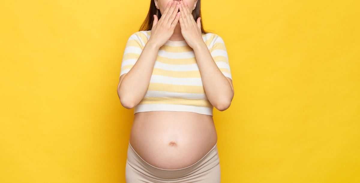 Shocked, astonished pregnant woman with bare belly wearing casual top isolated over yellow background screaming with amazement covering mouth with hands.