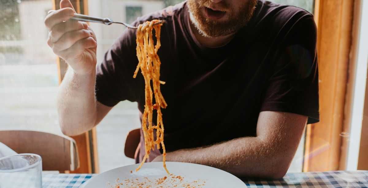 A man sits at a table, covered with a blue checkered tablecloth. He uses a fork to scoop up a mouthful of long spaghetti pasta. Meatballs rest on the large plate.