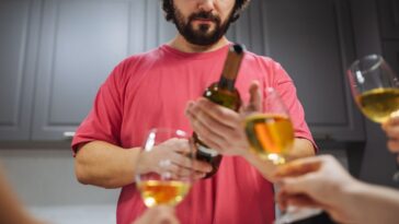 A man with a beard in a pink shirt is holding a bottle of wine while hands with wine glasses are extended to him. The background shows a kitchen scene. Suitable for gatherings, parties, and social events.