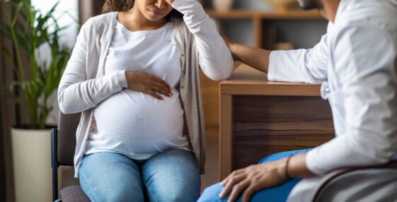 A pregnant young, woman feeling bad, sitting in a chair at a clinic, touching her big tummy and head, visiting doctor, gynecologist comforting crying expecting lady.