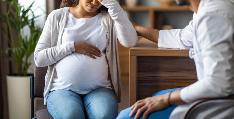 A pregnant young, woman feeling bad, sitting in a chair at a clinic, touching her big tummy and head, visiting doctor, gynecologist comforting crying expecting lady.